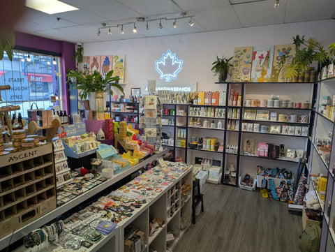 The Local Boom interior with shelves and counters displaying many colorful products, plants, wall art, and a glowing maple leaf sign on the back wall. The store has wood flooring, white walls, and mixed merchandise including packaged goods, books, and accessories.