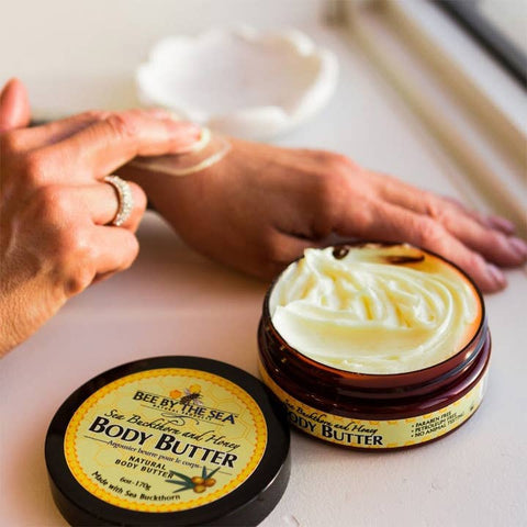 A person applying white body butter cream to their hand next to an open amber jar of Bee by the Sea body butter and its black lid on a light beige surface.